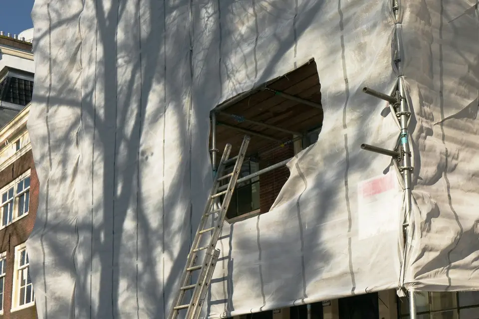 Een steiger met een ladder voor een gevel, ingepakt in wit doek waarop de schaduw van een boom valt