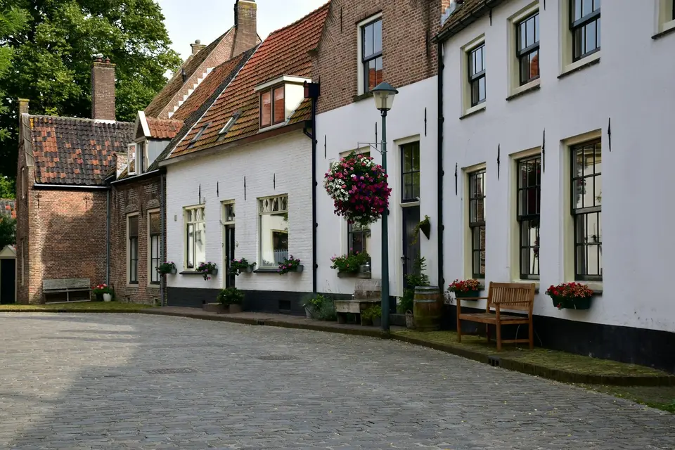 Een straat met klinkers waar witgepleisterde en bakstenen huizen naast elkaar staan, met bloembakken en een bankje