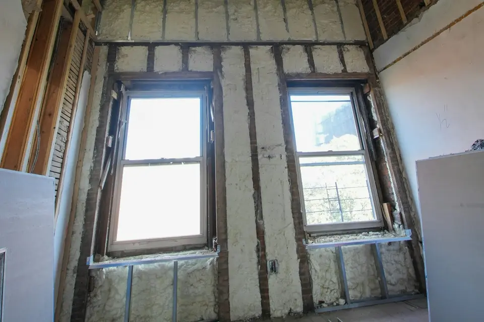 Een kamer in verbouwing met een frame van staal en hout tegen de buitenmuur, isolatie ertussen en twee ramen