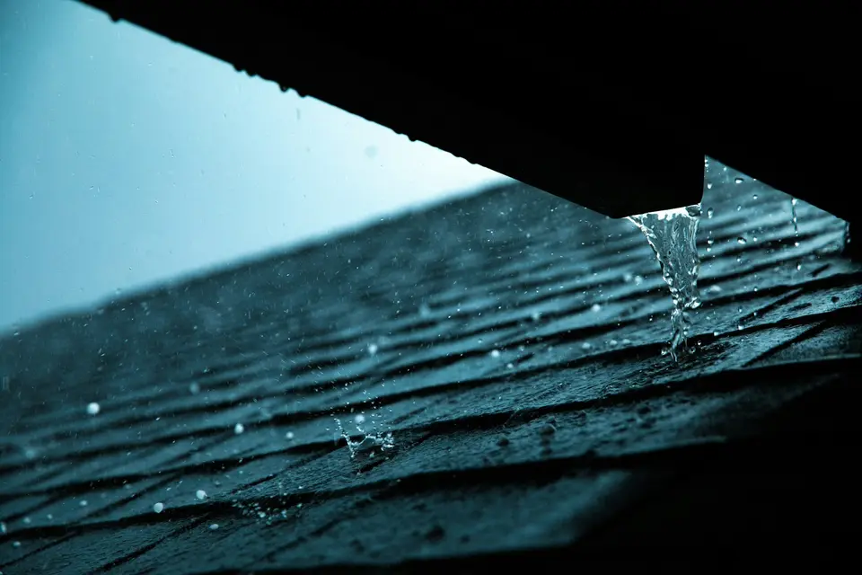 Regen loopt van een dakrand naar beneden, van onderaf gezien tegen een grijze lucht